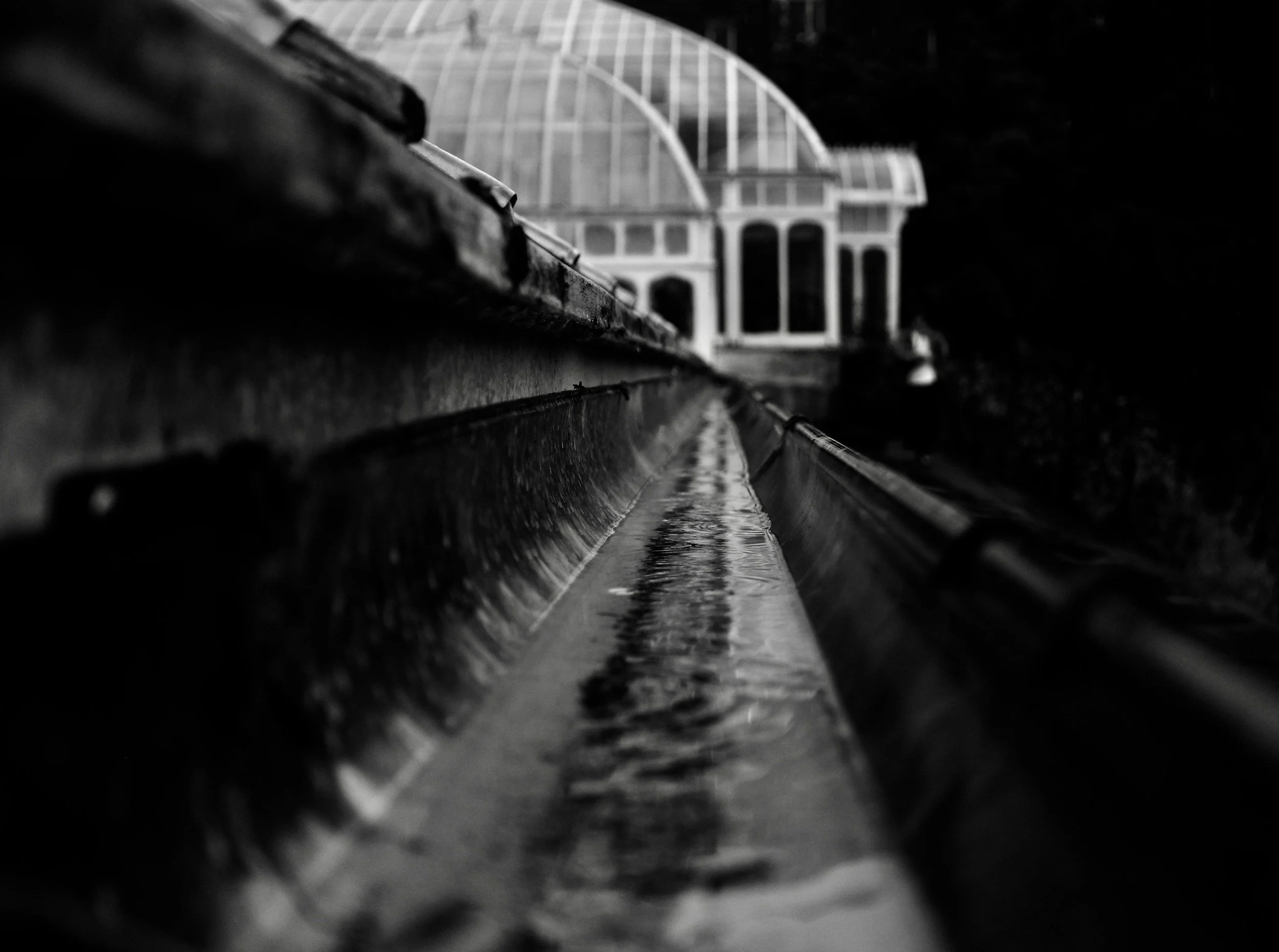 Black and white photo of a water channel or rain gutter with a greenhouse or conservatory in the background, seen from a low angle.