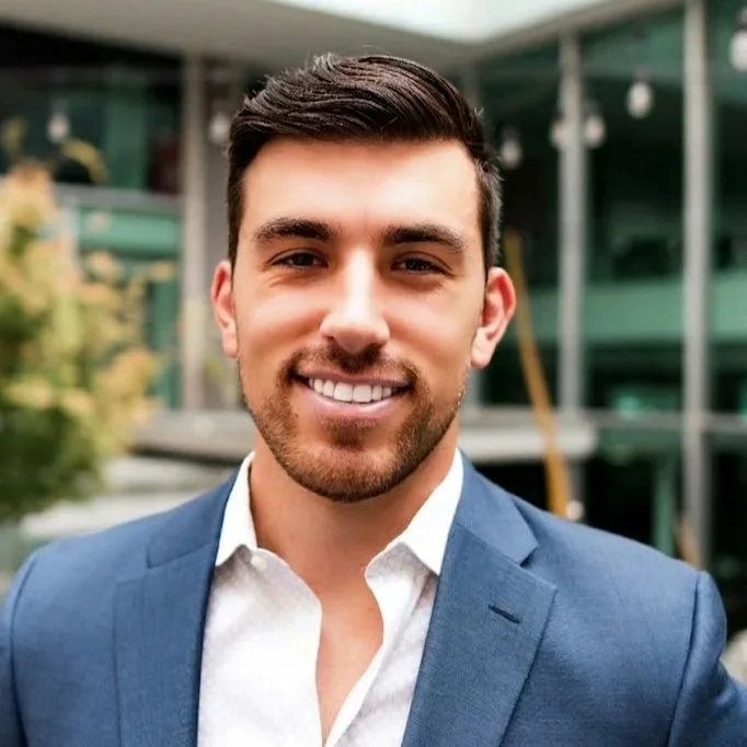 A smiling young man with dark hair and a beard, wearing a blue suit jacket and white shirt, standing outdoors with a modern building in the background.