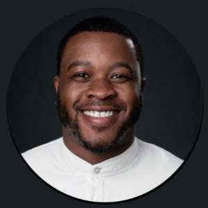 Close-up portrait of a smiling Black man with short black hair and a beard, wearing a white collared shirt, against a dark background.