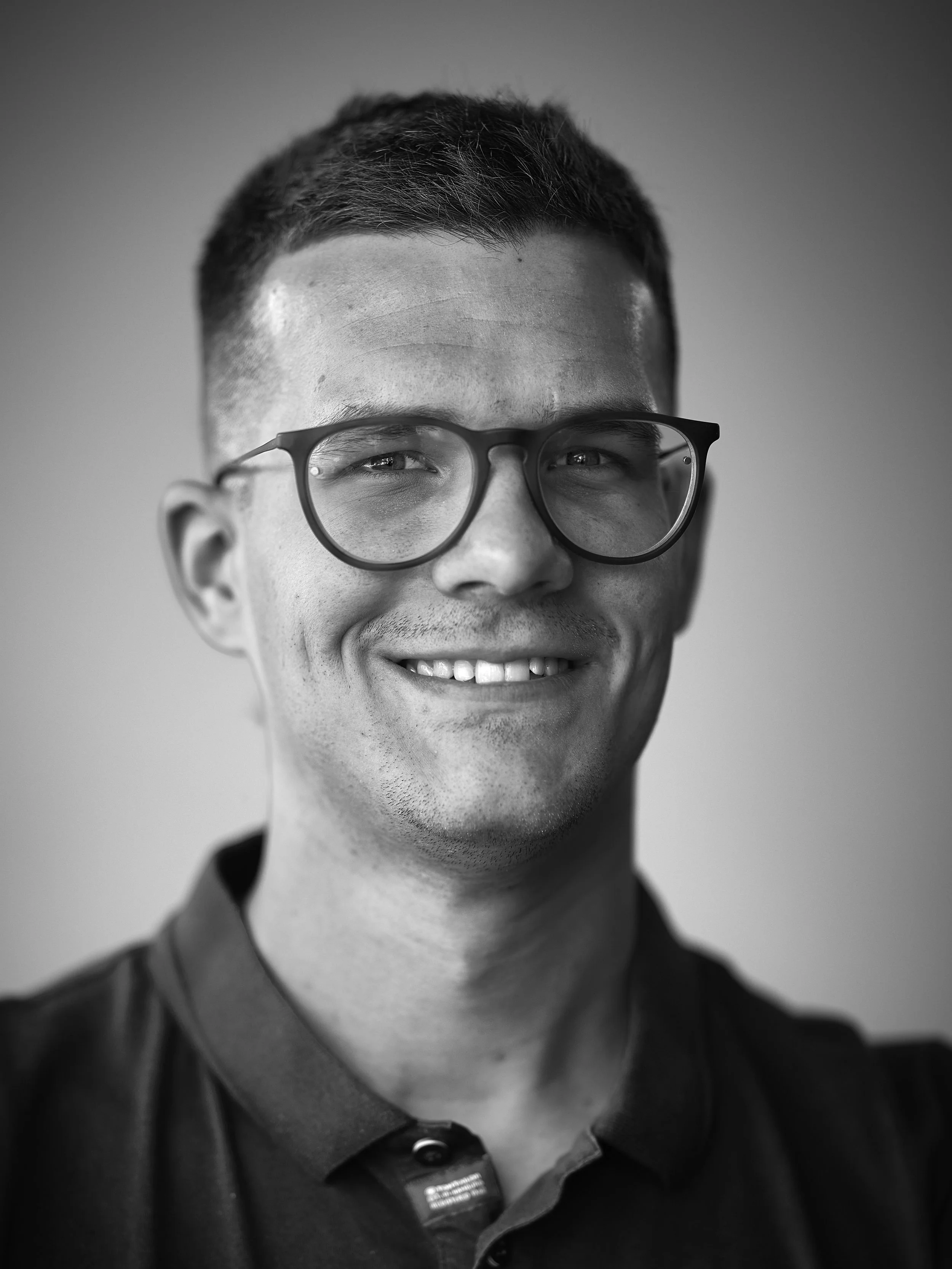 Close-up black and white portrait of a man wearing glasses and a collared shirt, smiling.
