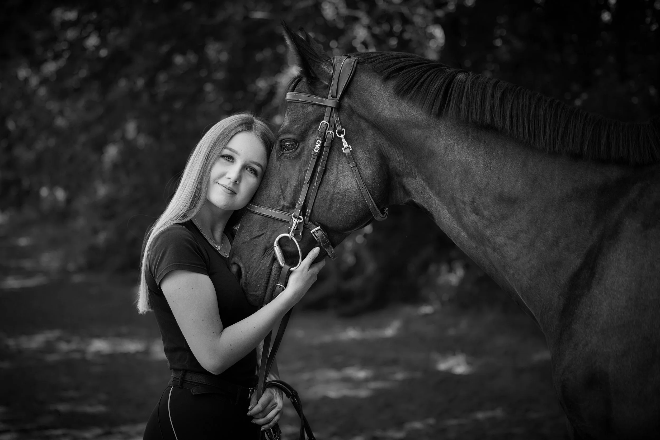 A woman with long hair and a dark shirt gently embraces the head of a dark horse, with her face close to the horse's face, in a natural outdoor setting.