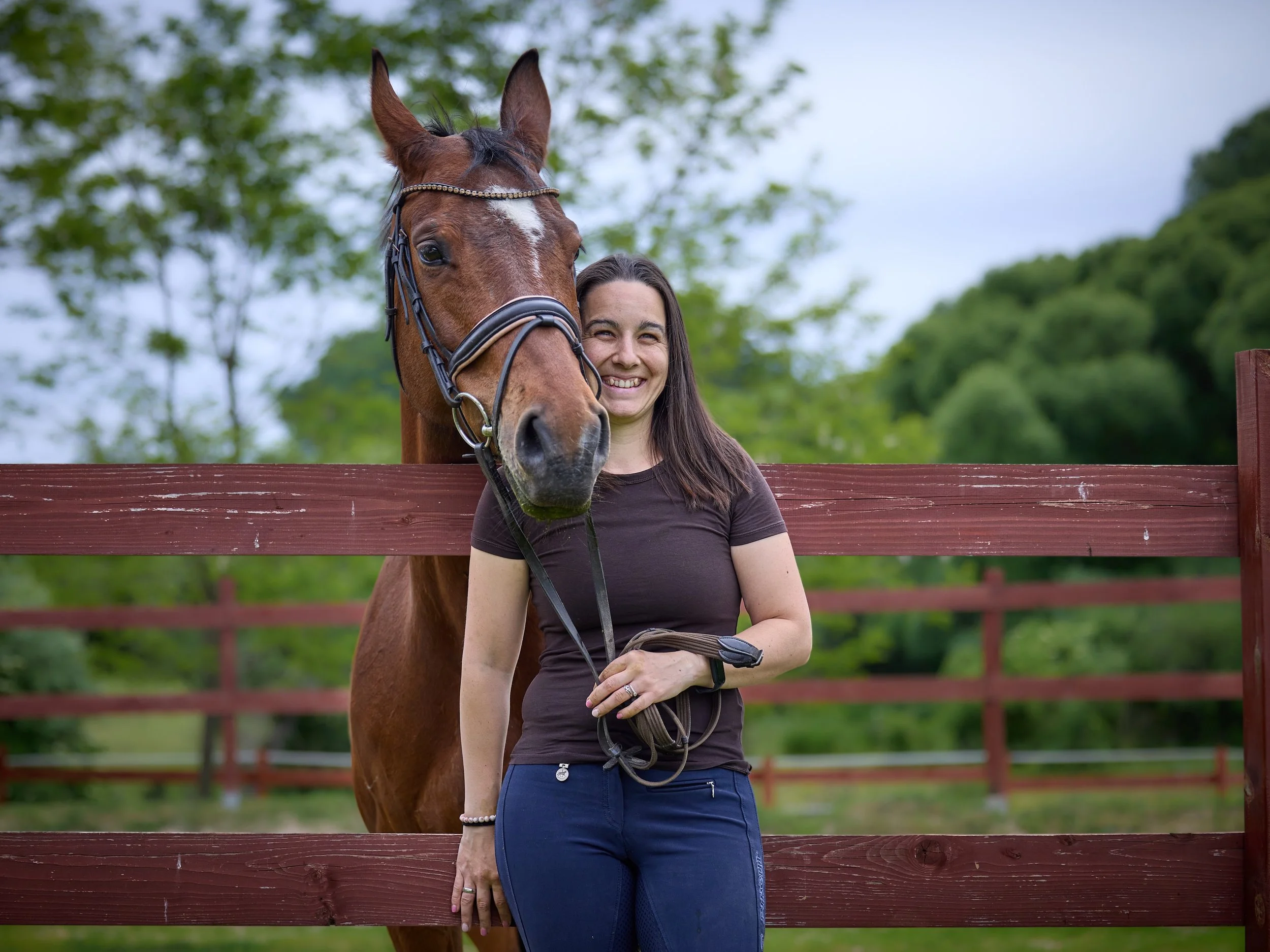 A woman with dark hair, wearing a black shirt, standing beside a brown horse with a white mark on its face, against a background of green trees and a cloudy sky, separated by a wooden fence.