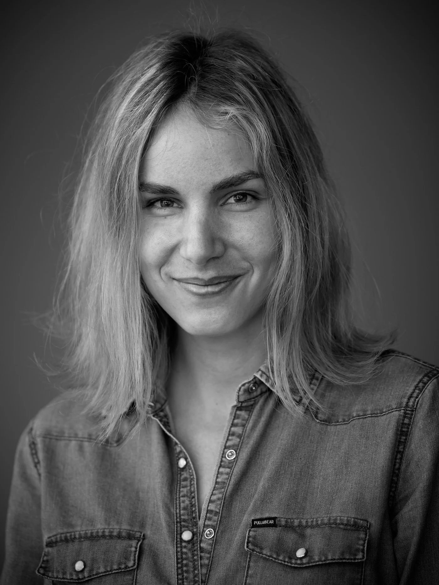 Black and white portrait of a woman with wavy hair, wearing a denim shirt with a small logo on the left pocket, smiling subtly.