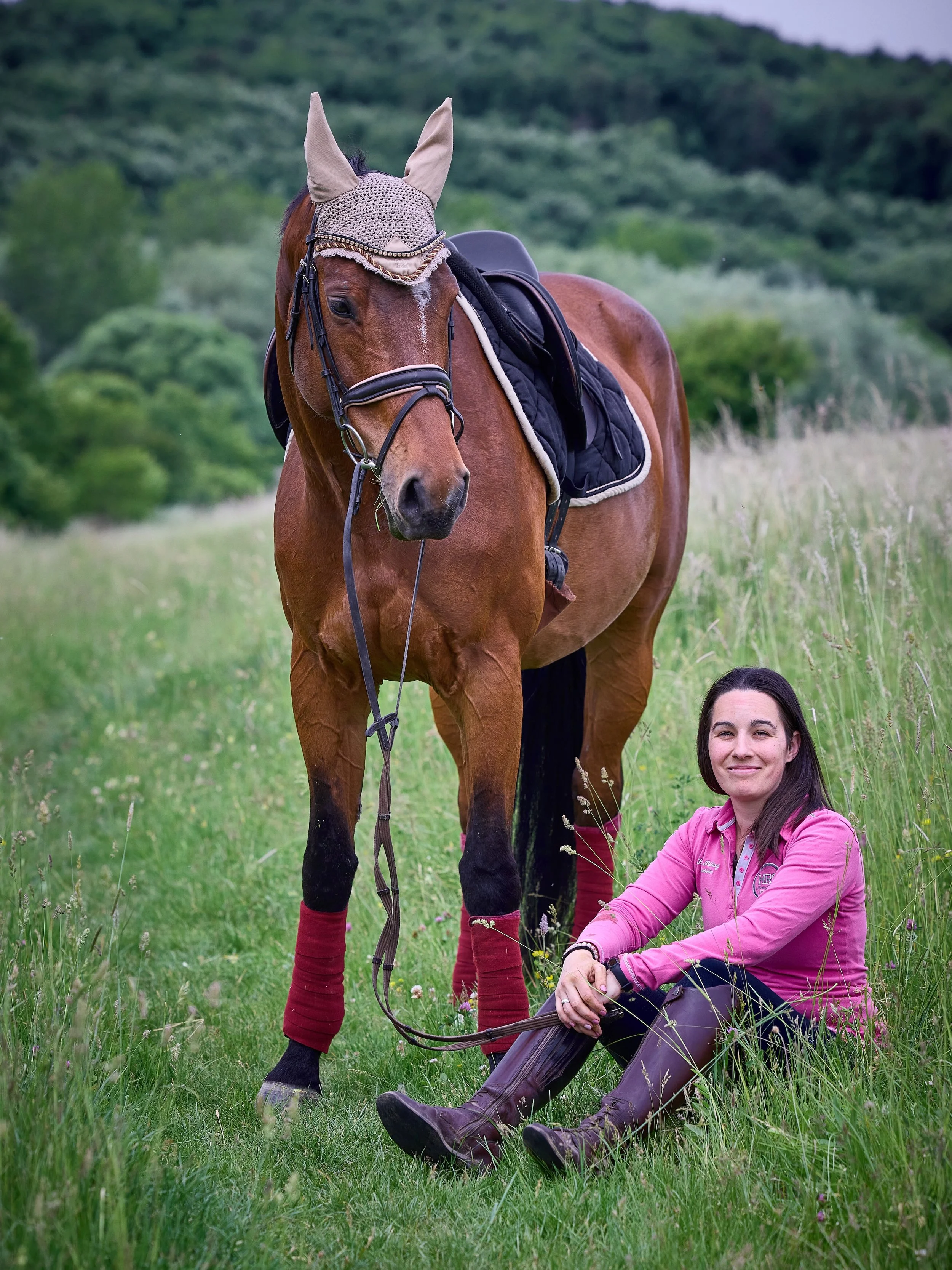 A woman with long dark hair sitting on grass next to a brown horse in a green field. The horse is wearing a crochet ear bonnet, a saddle, and red leg wraps. The woman is wearing a pink jacket and brown riding boots, smiling at the camera.
