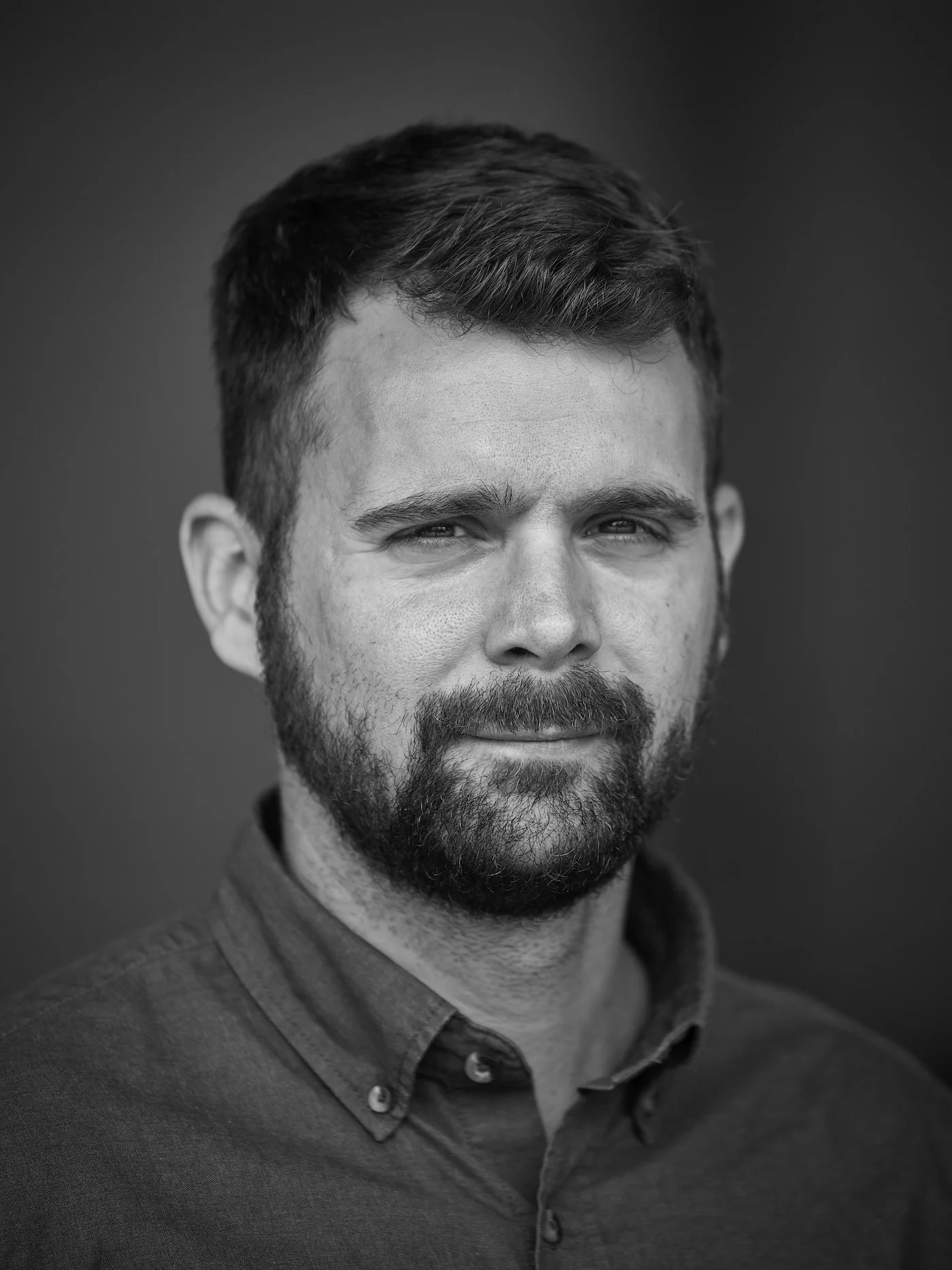 Black and white portrait of a man with short hair and a beard, wearing a button-up shirt with a collar, looking directly at the camera.