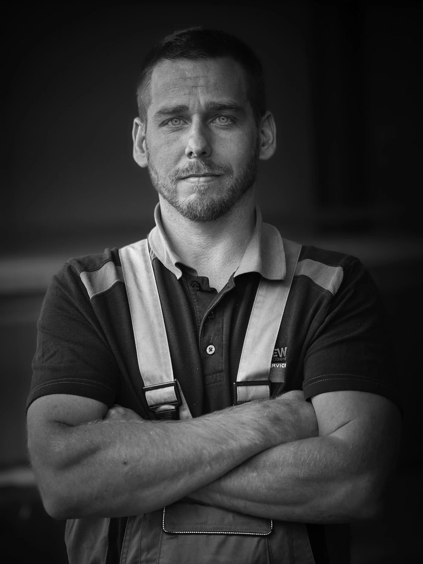 Black and white photo of a man with short hair and a beard, arms crossed, wearing a collared shirt and work vest, looking directly at the camera.
