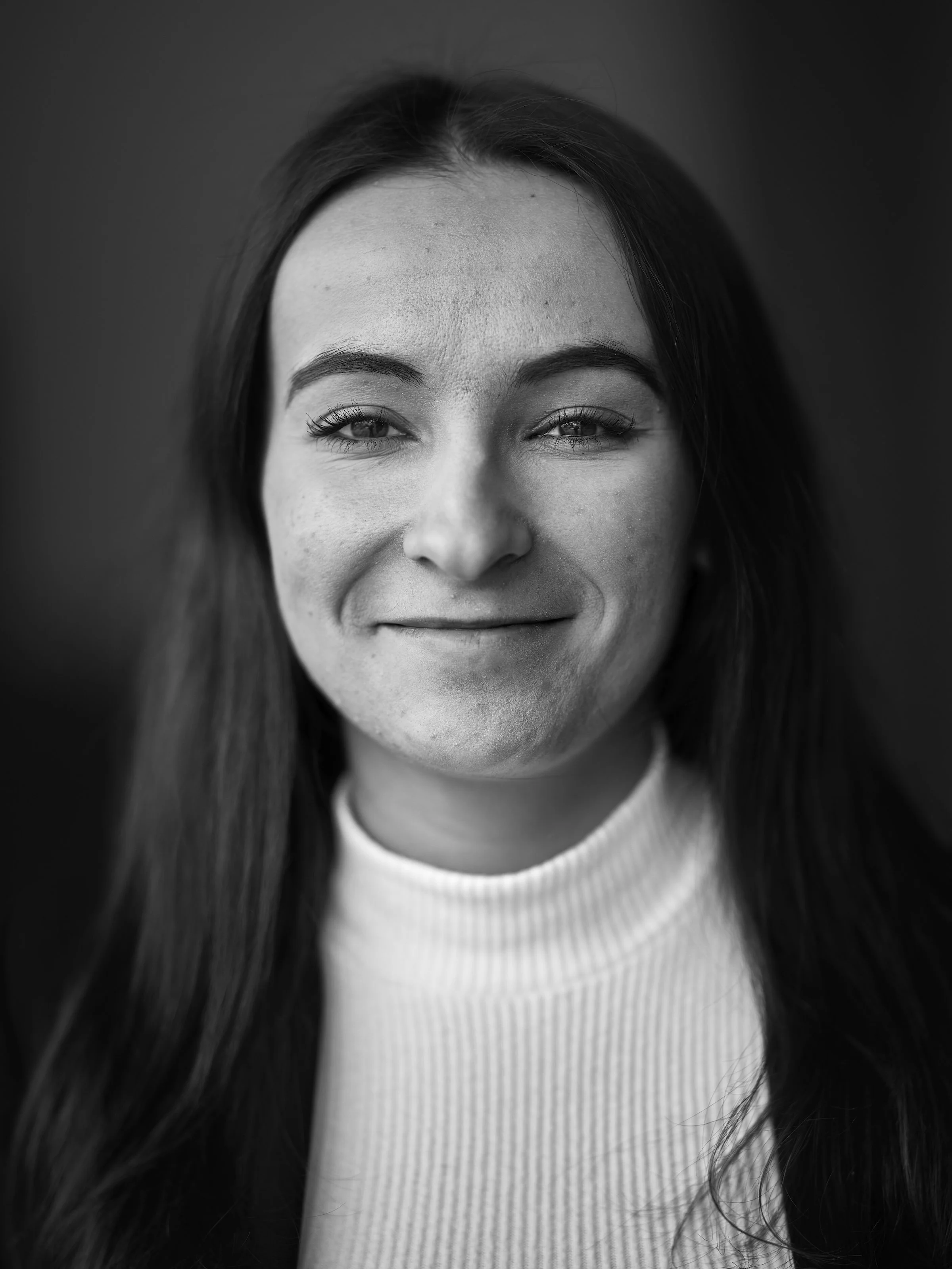 Close-up black and white photo of a woman with long dark hair, smiling slightly, wearing a light-colored ribbed turtleneck sweater.