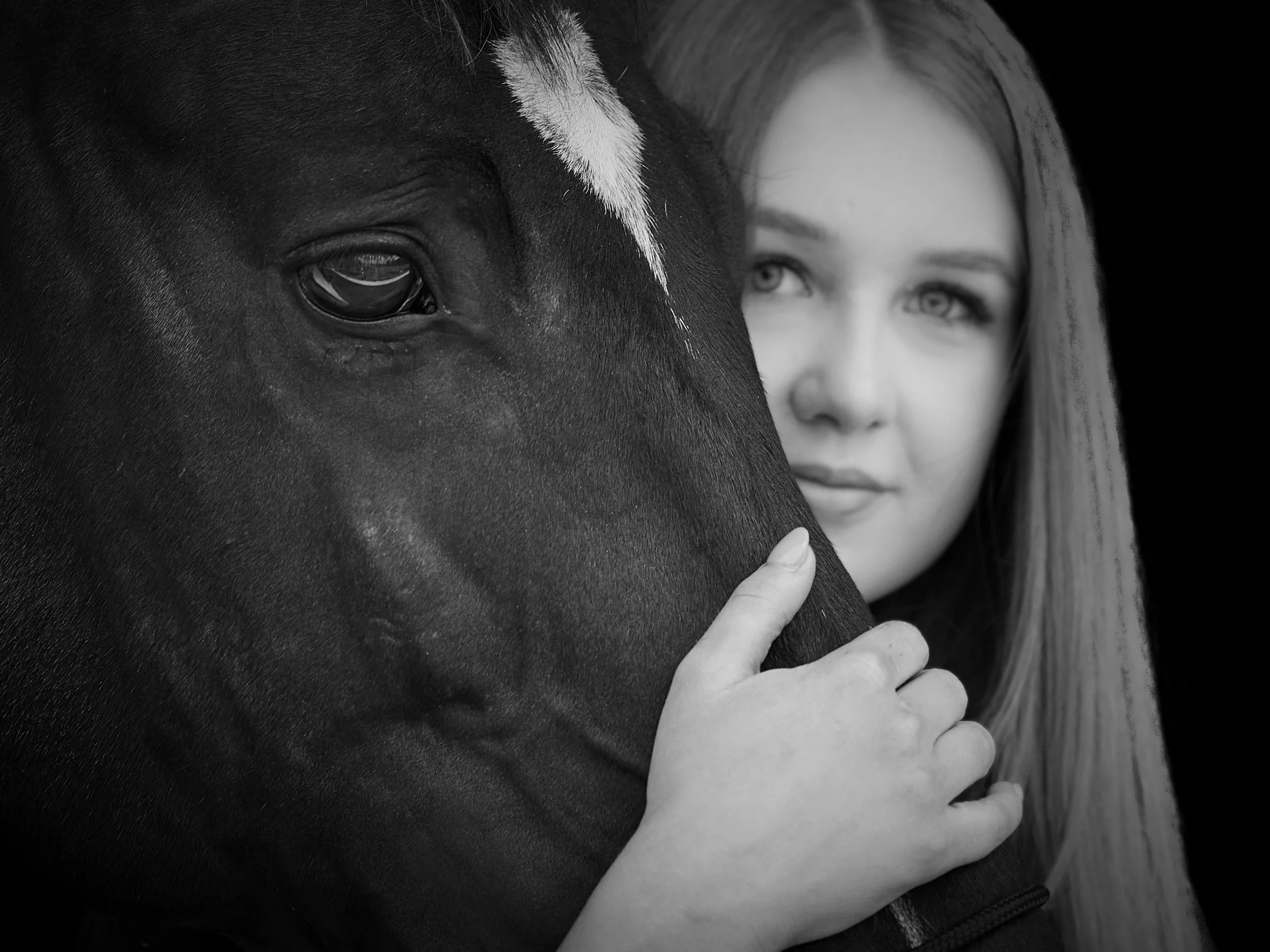 A woman with blonde hair and light eyes gently embracing a black horse with a white star on its forehead, both looking directly at the camera.