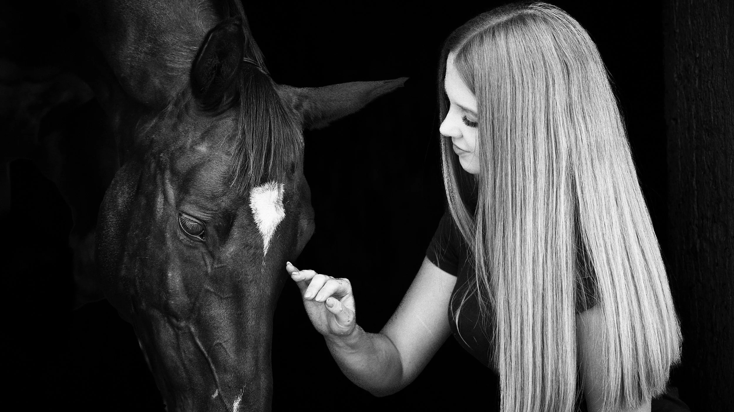 A young woman with long blonde hair gently touching the nose of a horse with a white star on its forehead, both in profile, against a dark background.