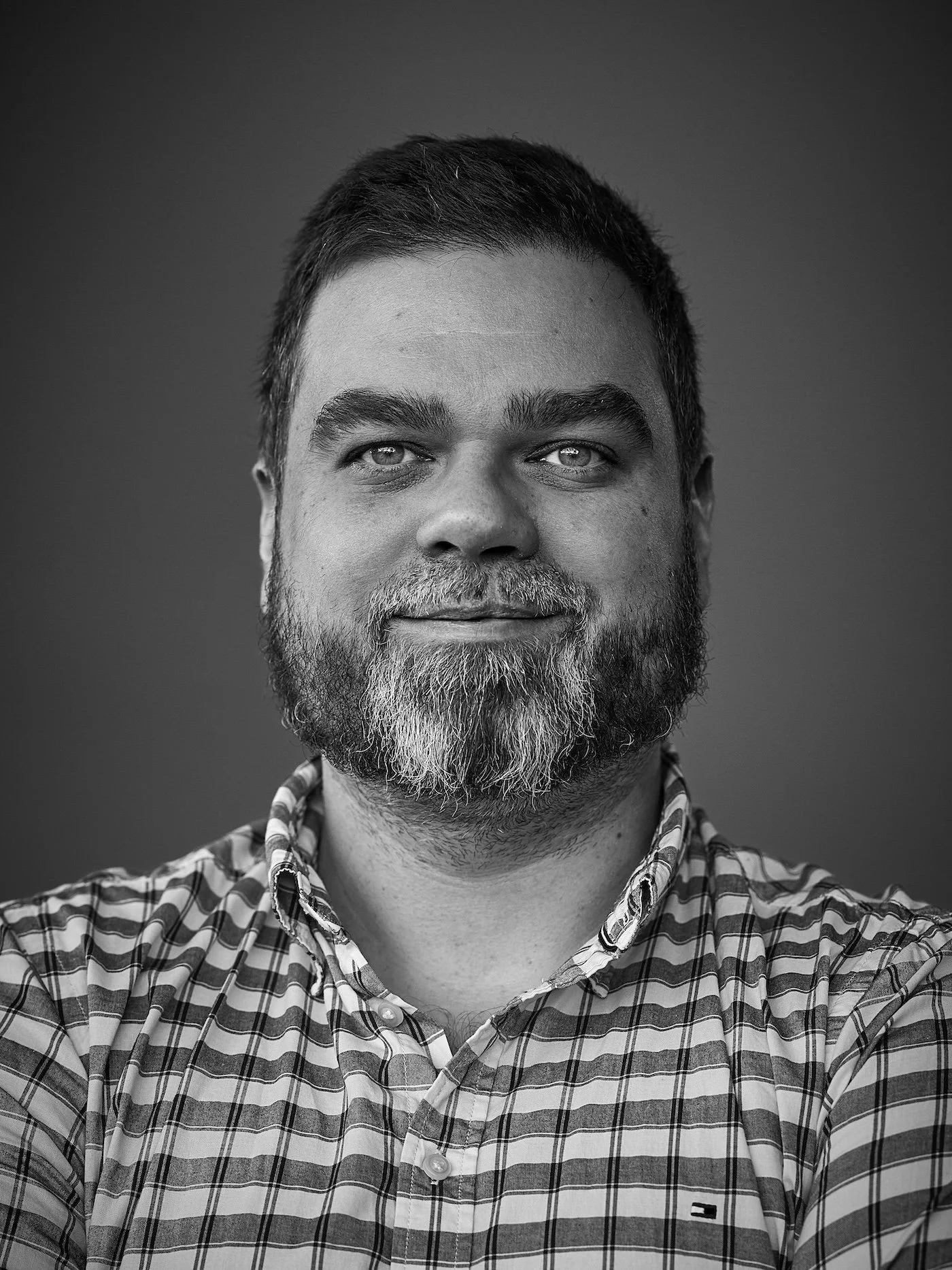 Black and white portrait of a man with short hair, beard, and mustache, wearing a checkered shirt with a small logo on it, smiling slightly, with arms crossed against a plain background.