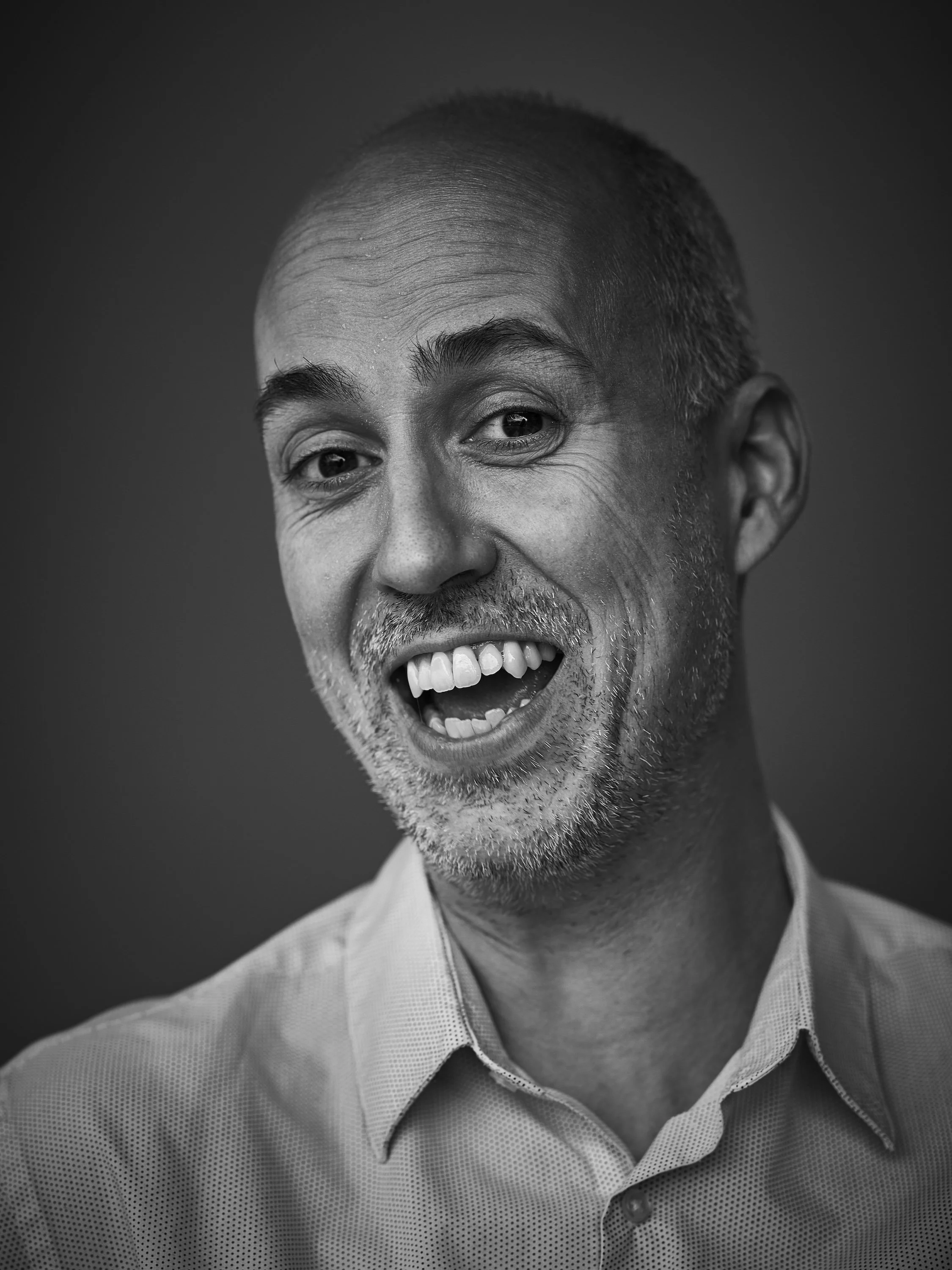 Black and white close-up portrait of a middle-aged man with short hair and a shaven face, smiling with his mouth open, wearing a collared shirt.