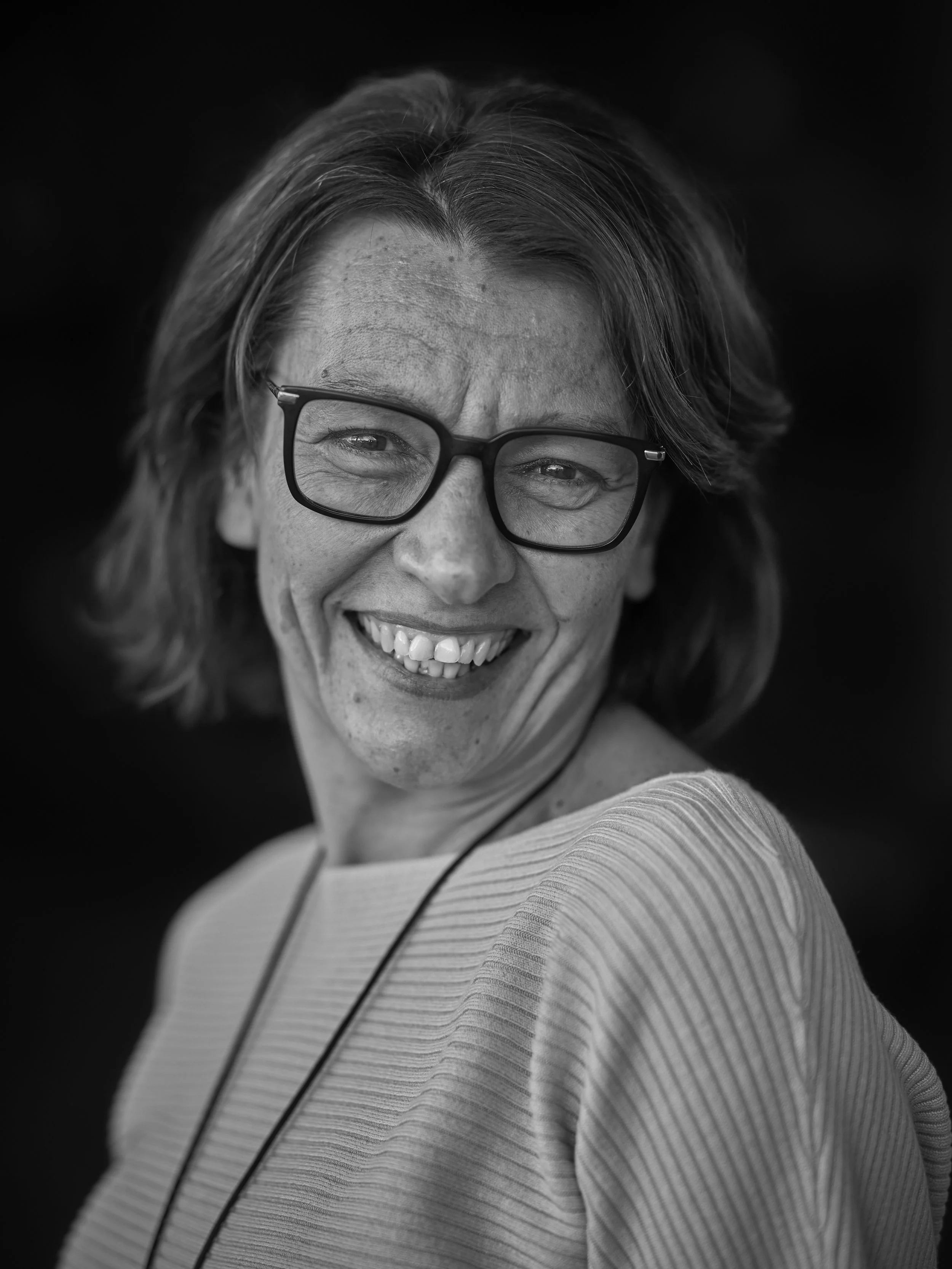 A smiling woman with glasses, shoulder-length hair, and wearing a striped top, against a dark background.