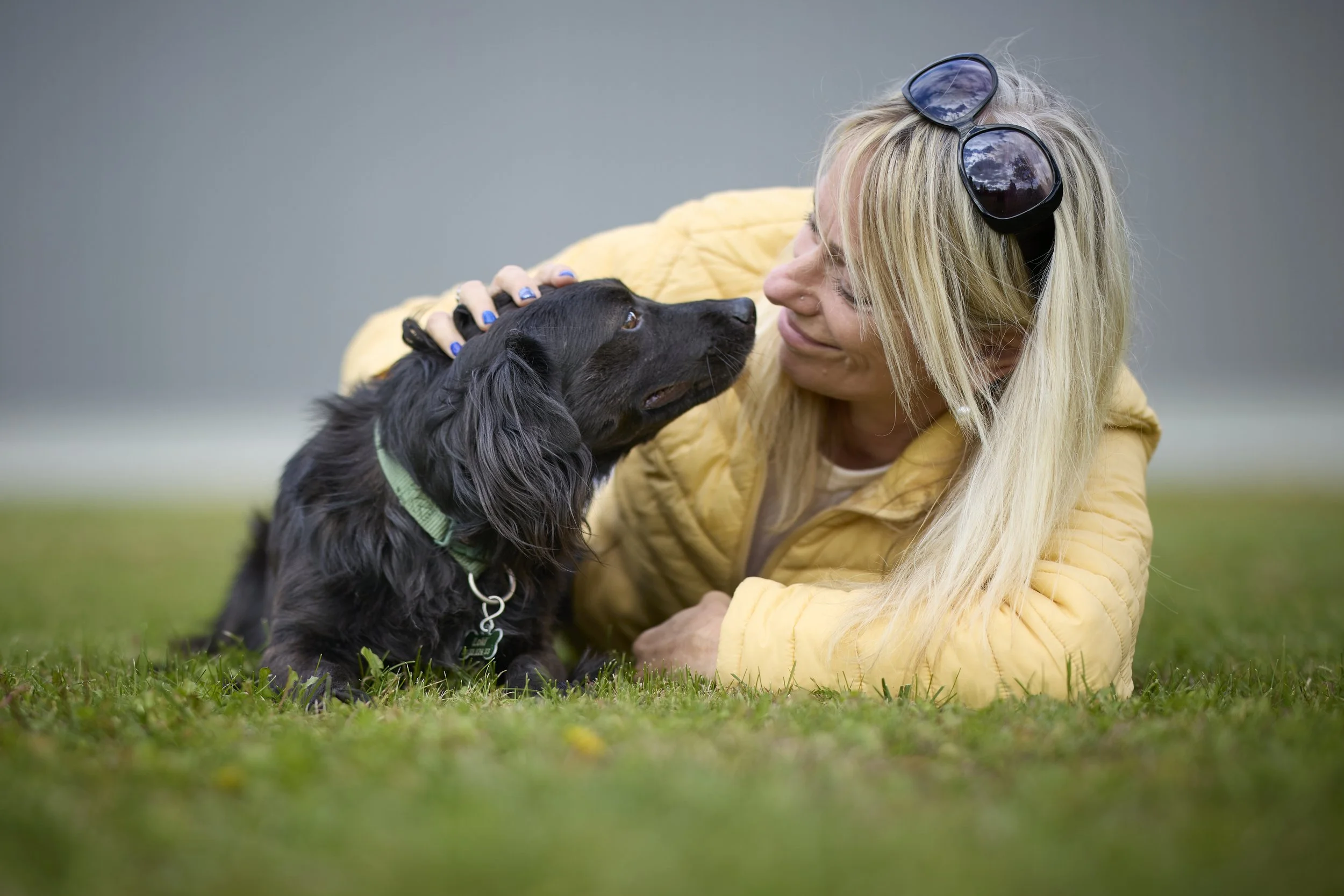 A woman with long blonde hair, wearing a yellow jacket and sunglasses on her head, gently holds a black dog with a green collar on the ground outdoors. They are making eye contact and appearing affectionate against a blurred green and gray background