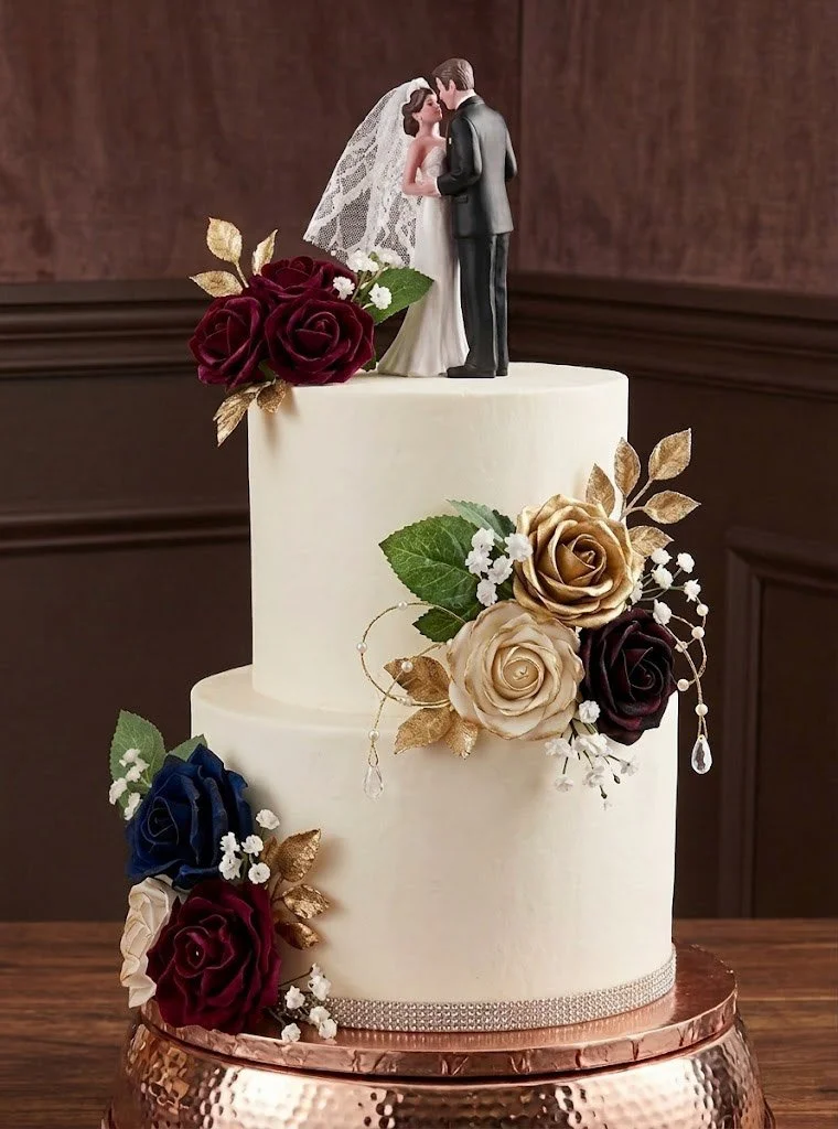 A wedding cake with a figurine of a bride and groom on top, decorated with burgundy, navy, cream, and gold roses, green leaves, and small white flowers.