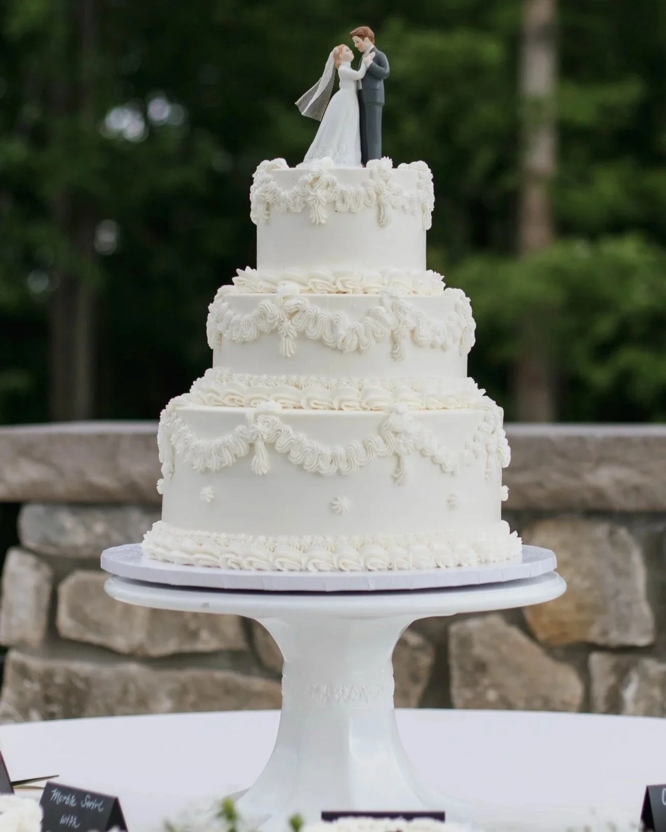 A three-tiered white vintage style wedding cake with decorative icing and a cake topper featuring a bride and groom figurine.