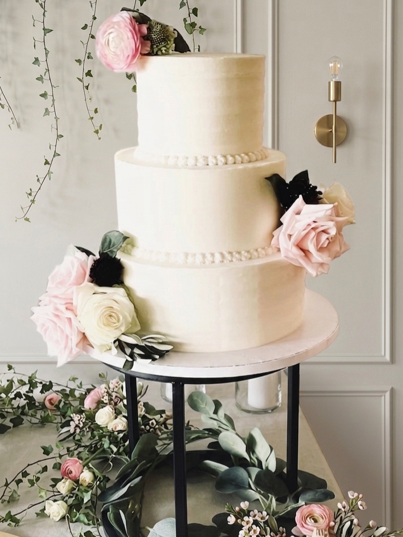 Three-tier vanilla Hazelnut cake decorated with pink and white flowers, displayed on a black stand, surrounded by greenery and floral arrangements.