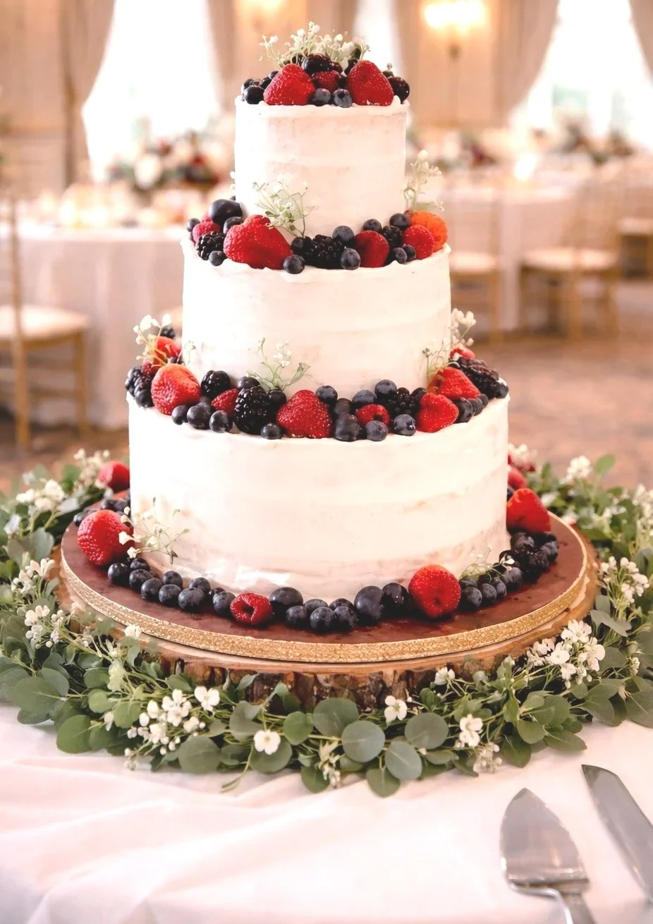 Three-tier white wedding cake decorated with strawberries, blueberries, blackberries, and greenery, set on a wooden base surrounded by floral greenery.