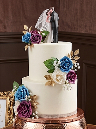 Wedding cake with white frosting, decorated with colorful blue, purple, and white roses, and topped with a bride and groom cake topper.