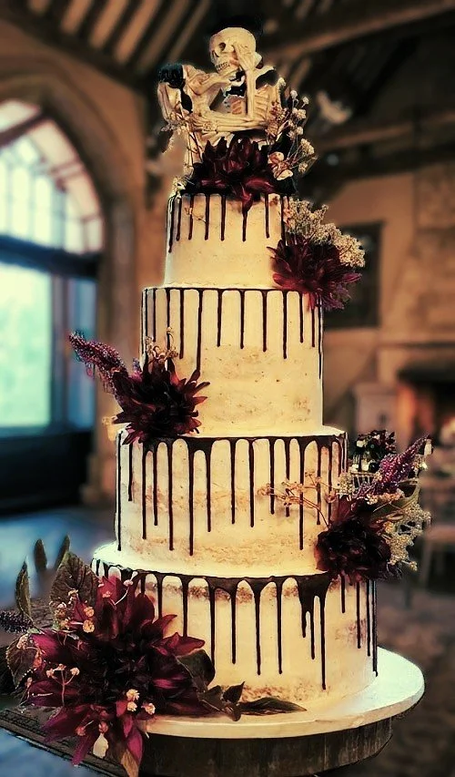 A tall, four-tier wedding cake decorated with dark red flowers and chocolate drips, topped with a skull and bones decoration.