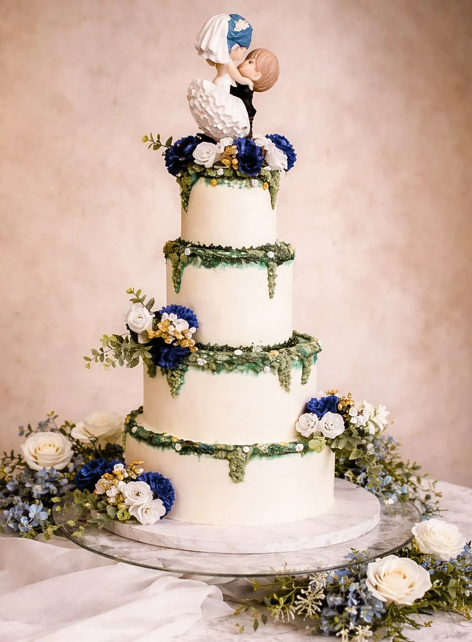 A multi-tiered wedding cake decorated with white, blue, and gold flowers, and topped with a figurine of a bride and groom kissing.