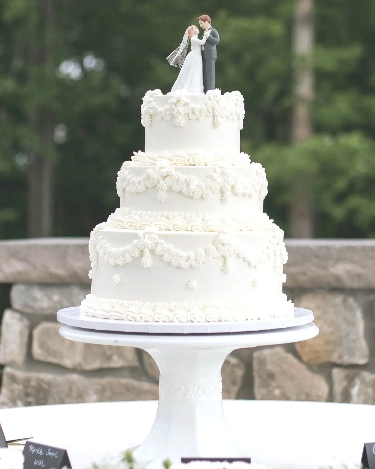 A four-tier white vintage wedding cake with ornate icing decorations and a bride and groom figurine on top, set outdoors with a stone wall and green trees in the background.