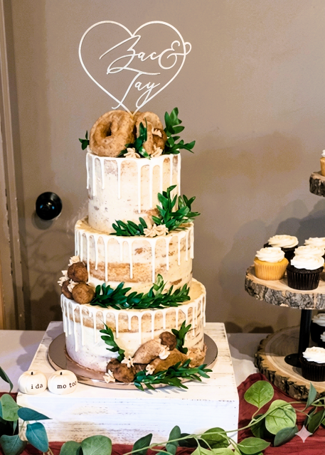 Three-tiered wedding cake with white drip icing, greenery decorations, and donuts on top. 