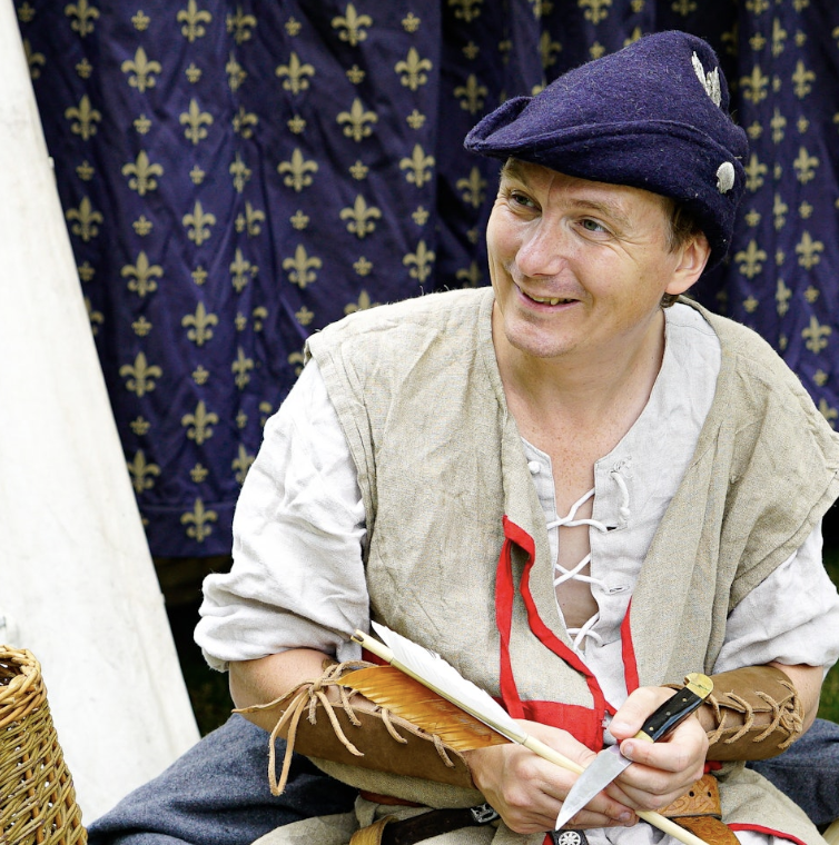 A man dressed in medieval costume including a blue cap, beige vest, and long-sleeved shirt, smiling while sitting outdoors with a fabric backdrop featuring a gold fleur-de-lis pattern.