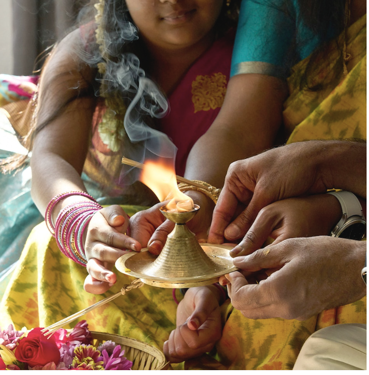 A woman wearing colorful traditional Indian attire lighting a small brass oil lamp with flickering flames, smoke rising from the wick, and flowers nearby.