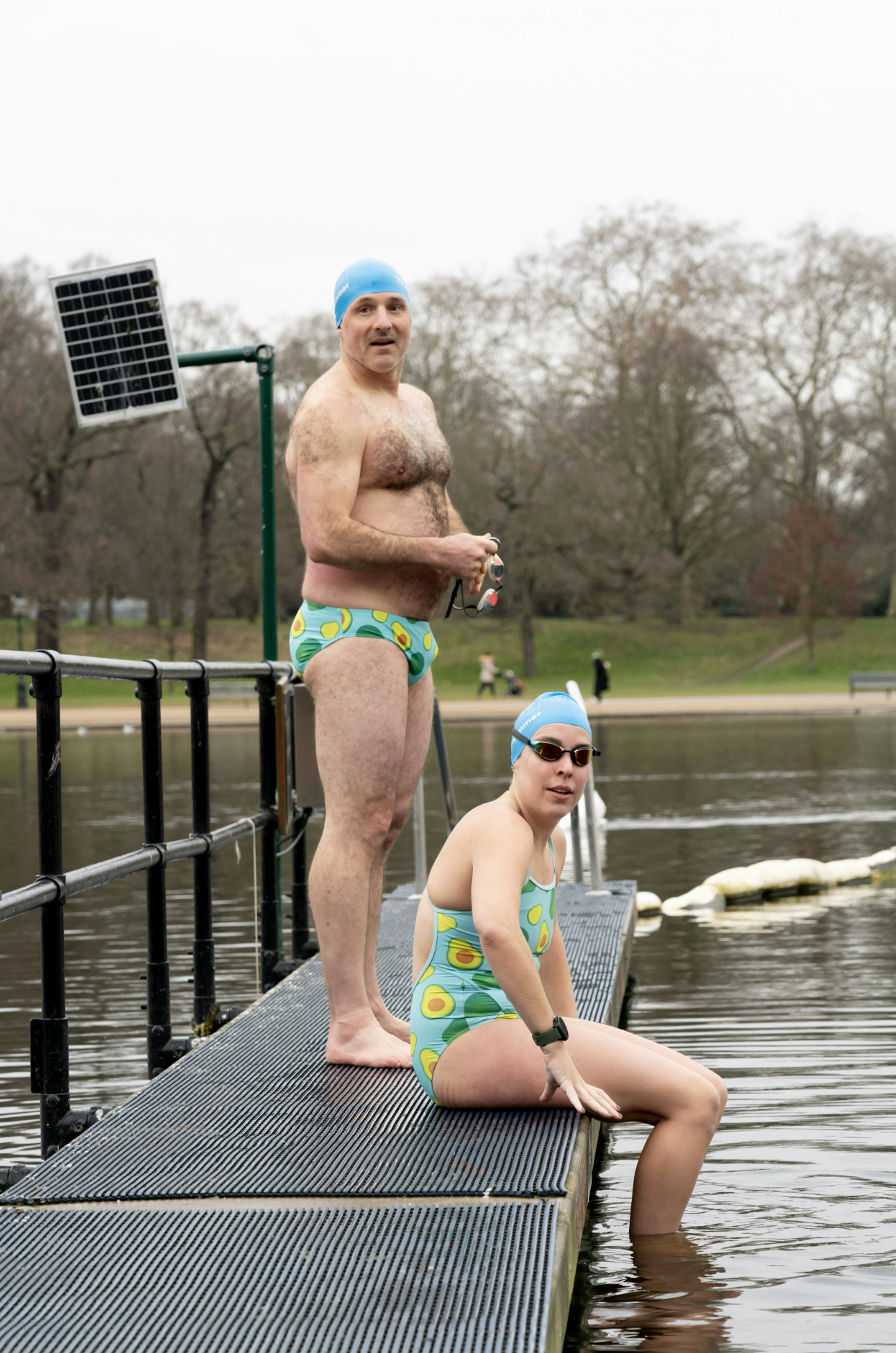 A man and woman in vintage-style swimsuits with avocado patterns, wearing swim caps and goggles, at a park lake. The woman is sitting on the dock with her feet in the water, while the man is standing behind her, holding goggles.