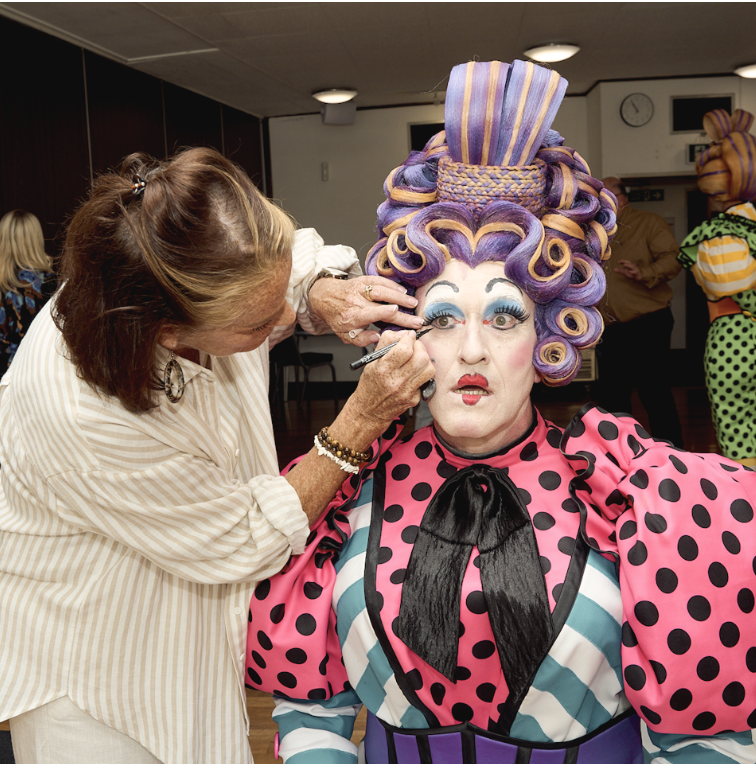 Makeup artist applying makeup on a drag queen with elaborate curly purple wig, bold makeup, and colorful polka-dot costume.