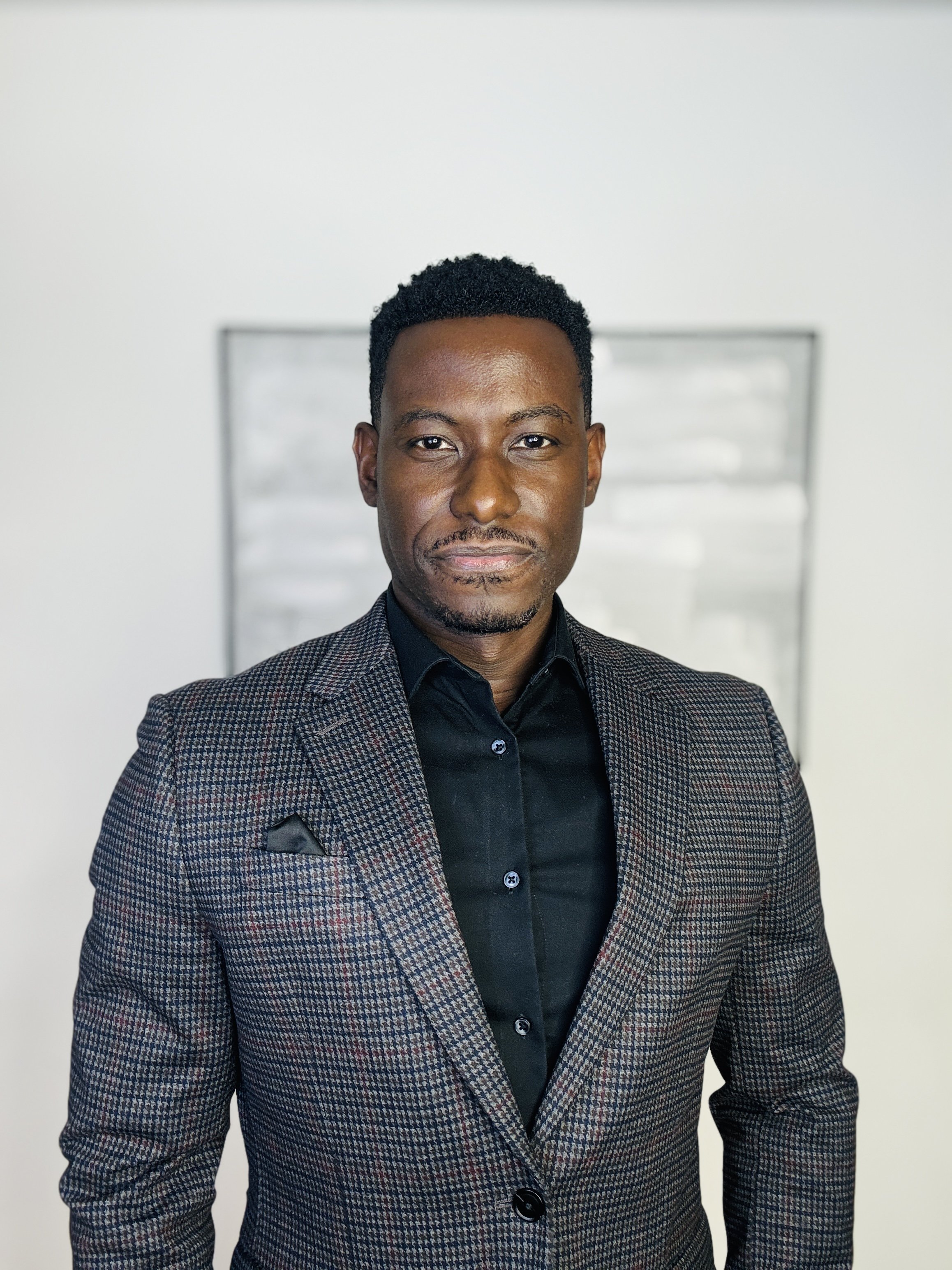 A professional man wearing a dark patterned suit and black shirt, standing against a plain light background.