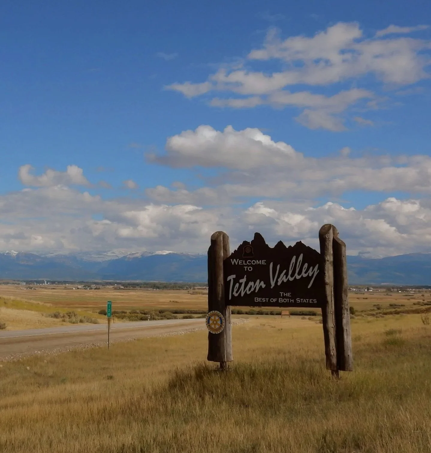 A welcome sign for Teton Valley with mountains in the background and a partly cloudy sky.