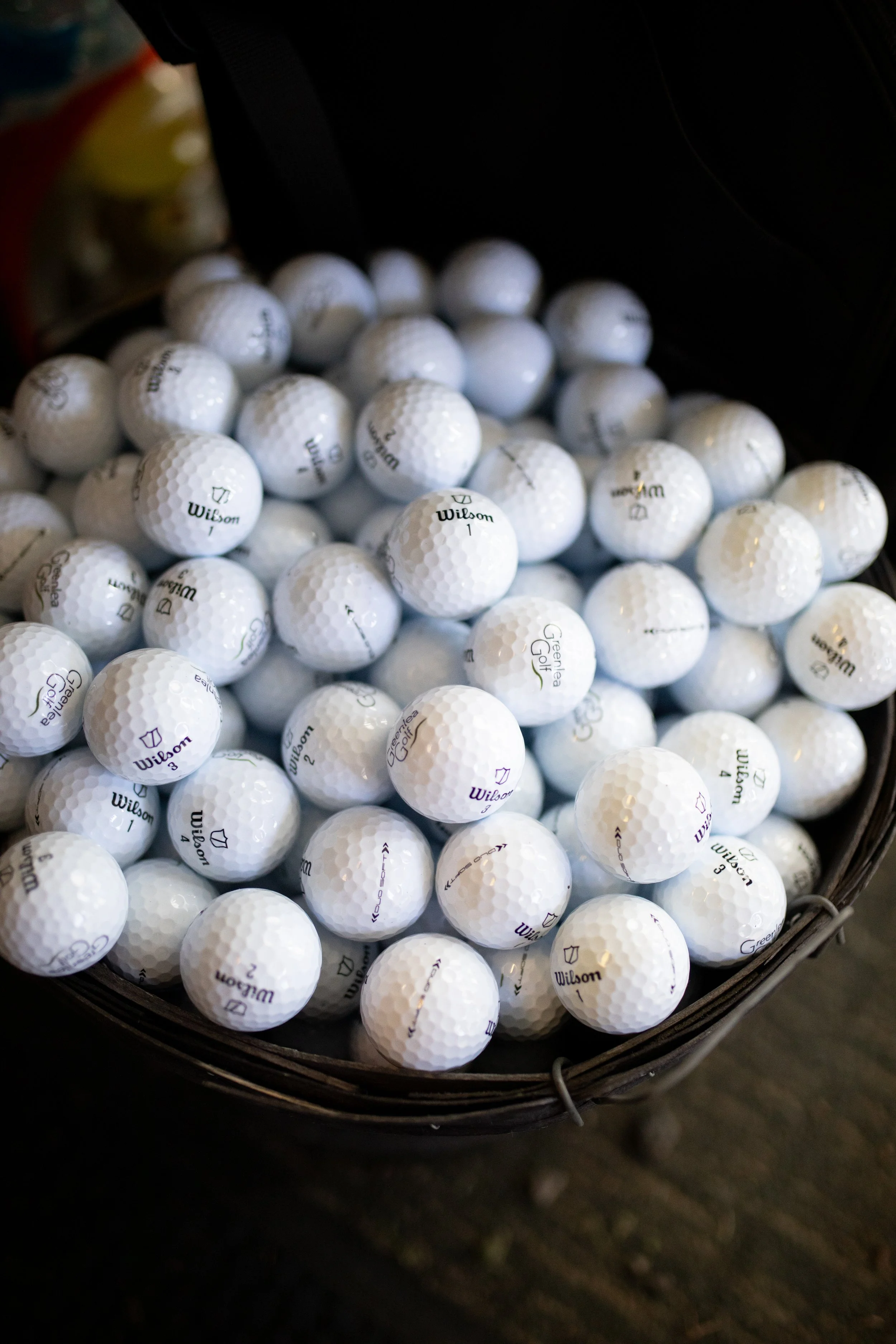 Basket of white golf balls with brand logos, including Wilson and Greenea Golf, on a dark background.
