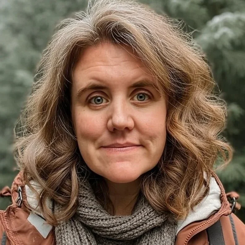 A woman with curly, shoulder-length brown hair and blue eyes looking at the camera outdoors, wearing a brown jacket and a gray scarf.