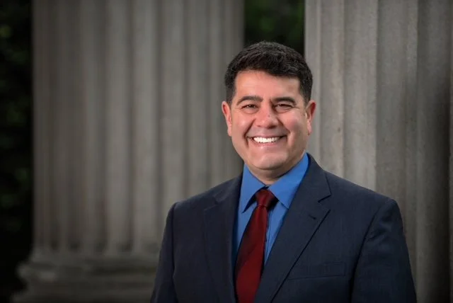 Neil Horible in a dark suit, blue shirt, and red tie standing outdoors in front of large stone columns, smiling at the camera.