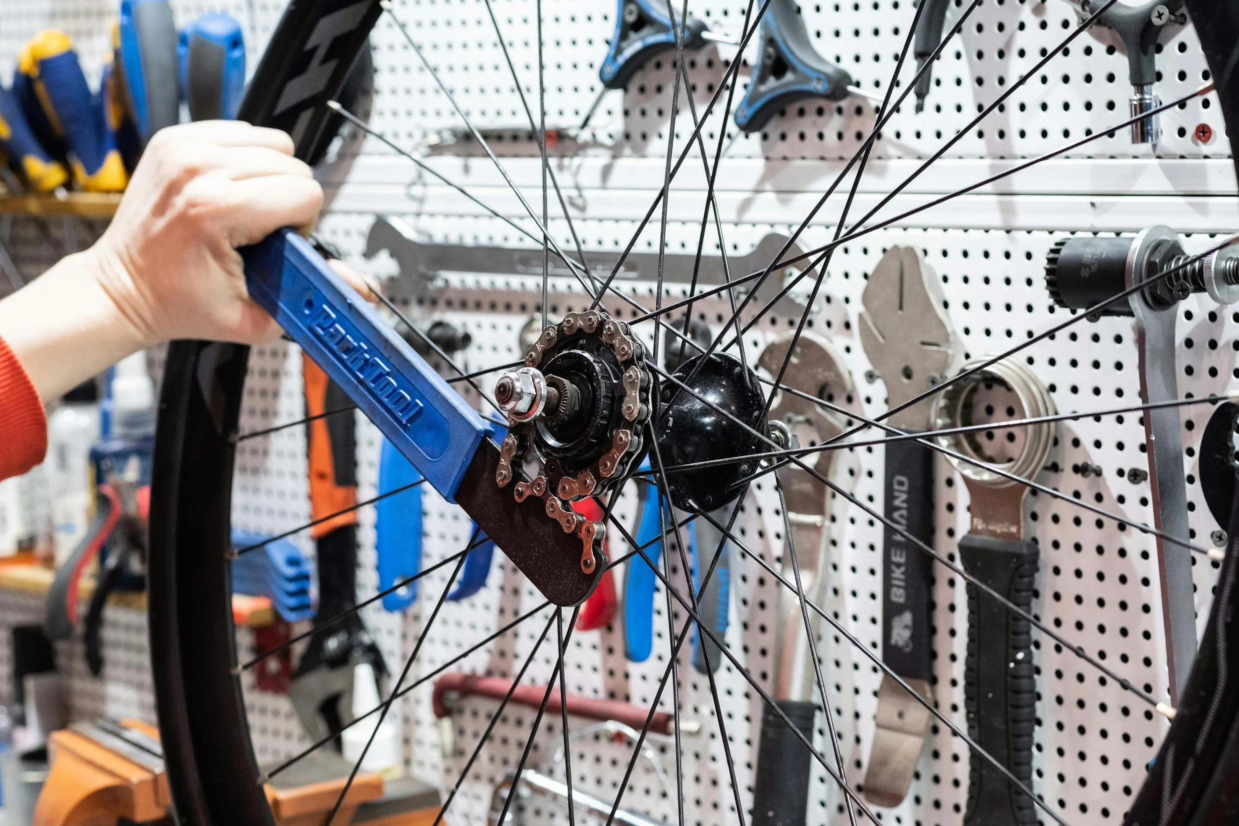 A person adjusting the gears of a bicycle wheel in a workshop, with various tools hanging on a pegboard in the background.