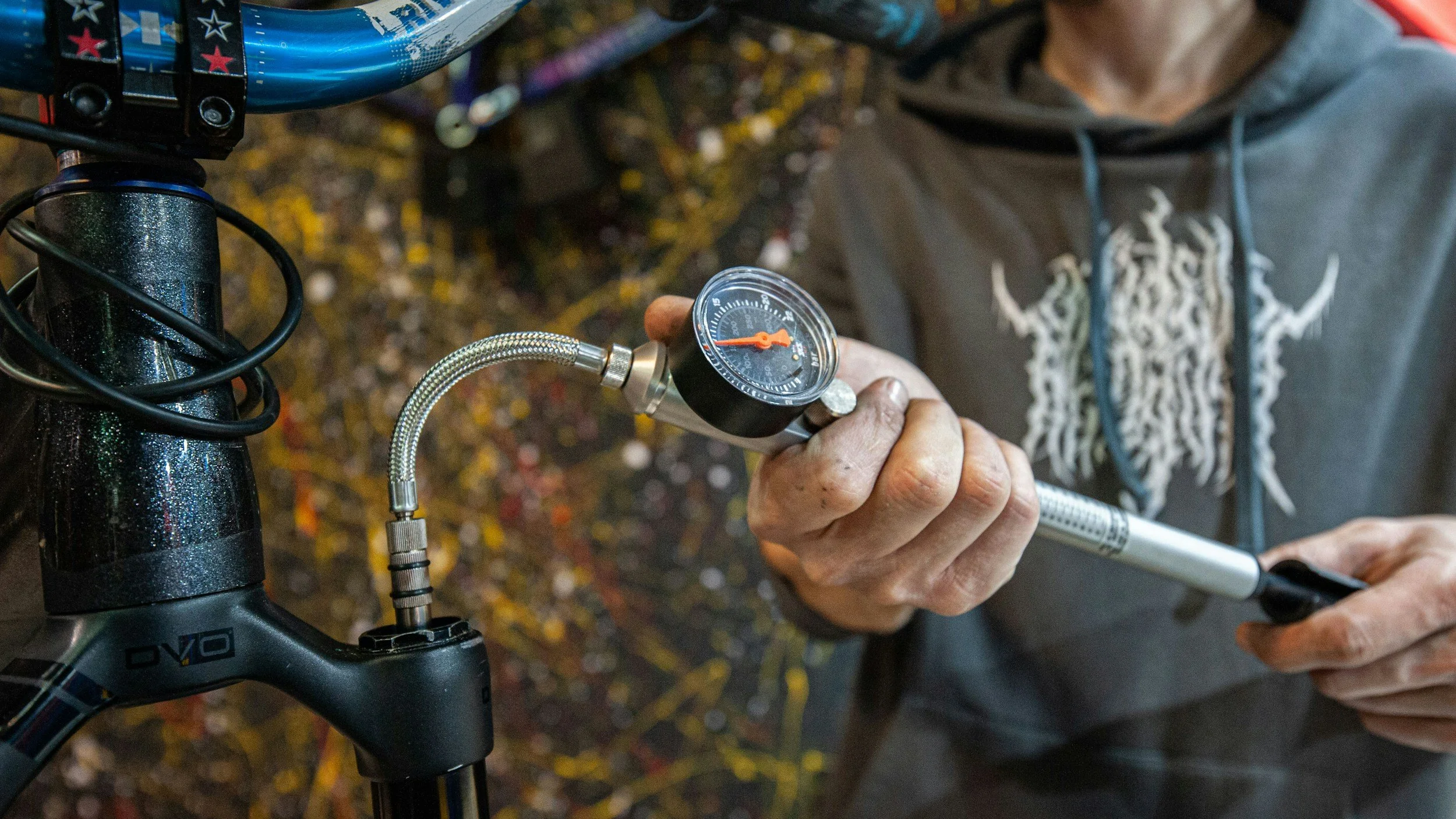 A person holding a pump gauge attached to a bicycle tire, with a black bicycle in the foreground and a colorful ground surface beneath.