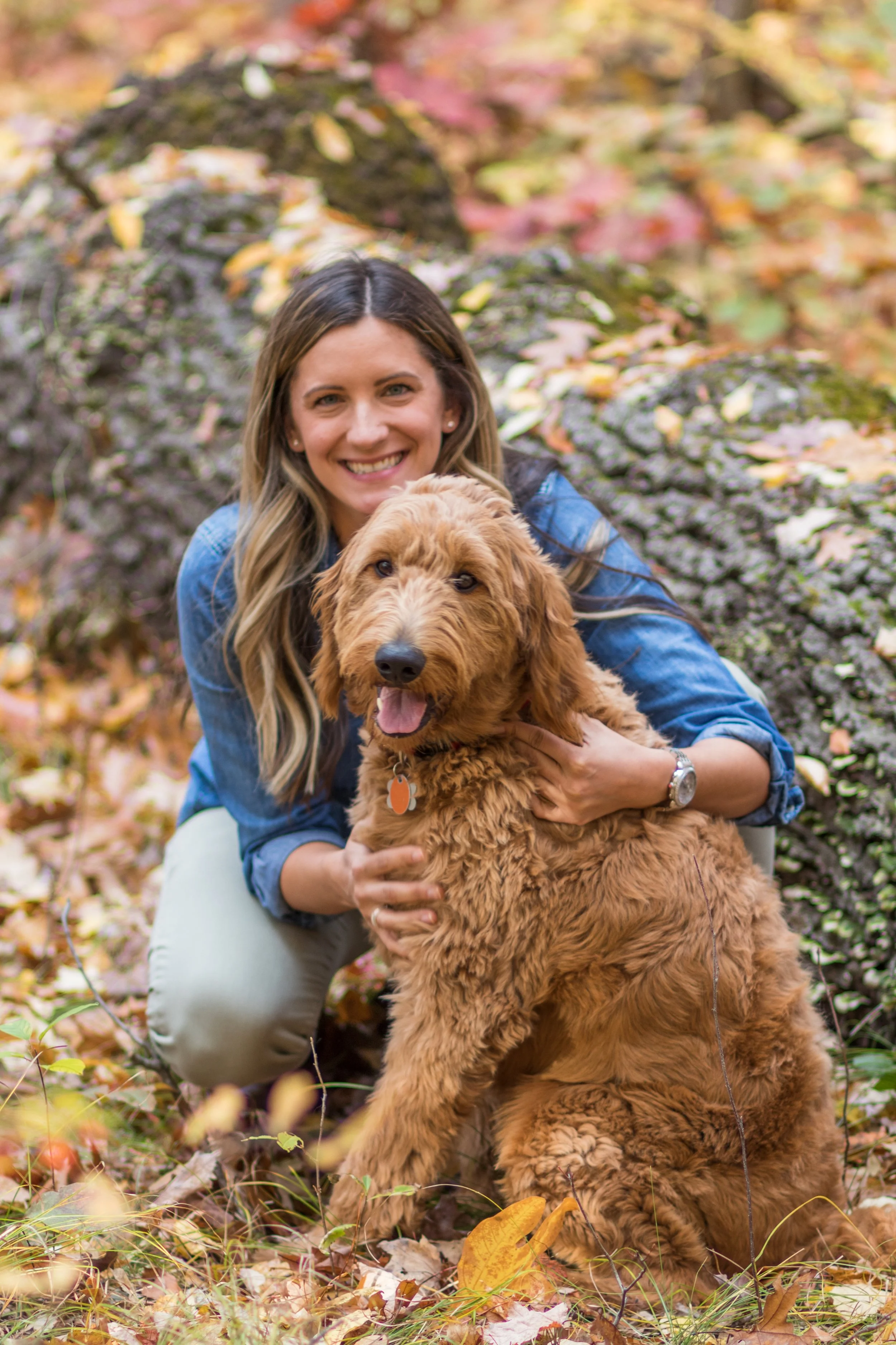A woman with long, wavy hair and a blue jacket smiling while holding a large, fluffy, golden retriever puppy in an outdoor setting surrounded by autumn leaves.