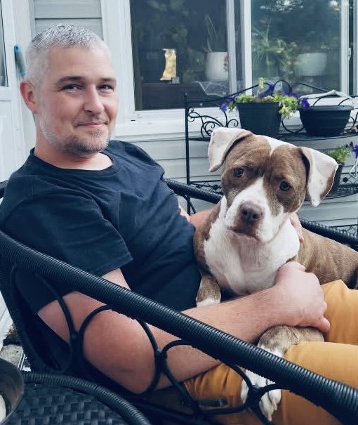 A man with short gray hair and a beard sitting on a black outdoor chair, holding a brown and white dog on his lap on a porch with a white house and potted plants in the background.