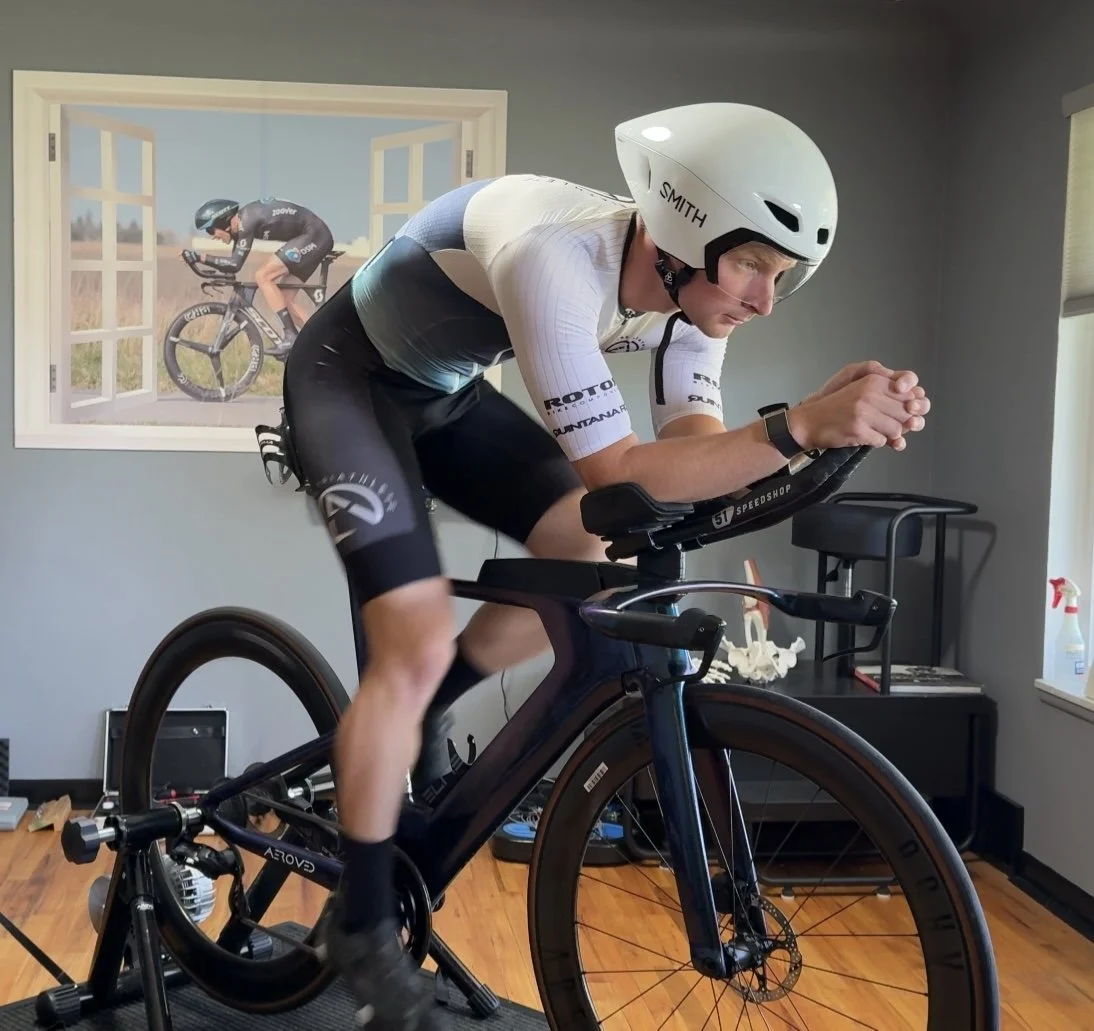 Man wearing a white helmet and cycling gear on a stationary indoor bike, with a window and a poster of a cyclist on the wall.