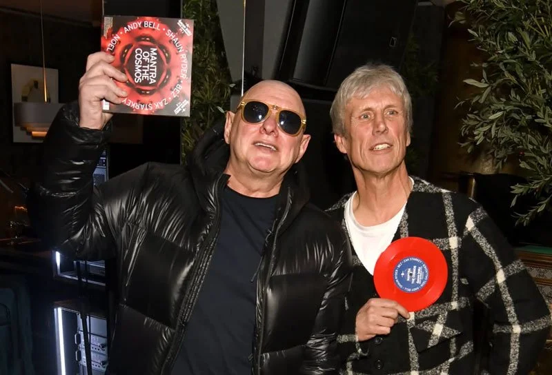 Two men posing indoors, one holding a vinyl record and the other holding a red frisbee.