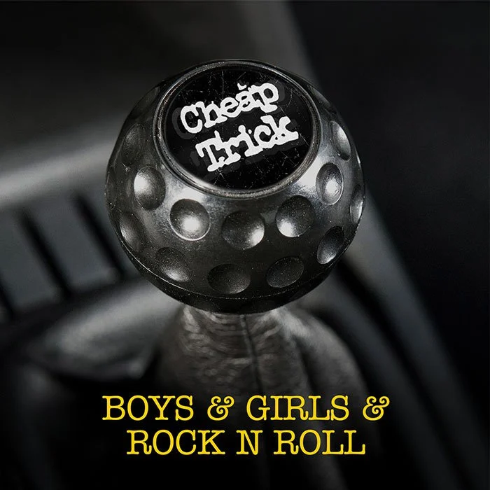 A gear shift knob with a black circular top displaying the words "Cheap Trick" in white text, and the base of the knob covered in a metallic, dimpled design. Below, yellow text reads "BOYS & GIRLS & ROCK N ROLL".