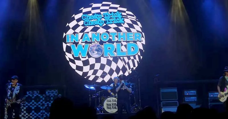 A band performing on stage with a large circular graphic behind them that reads 'Chicano Thak Chacho Thak' and 'In Another World'.