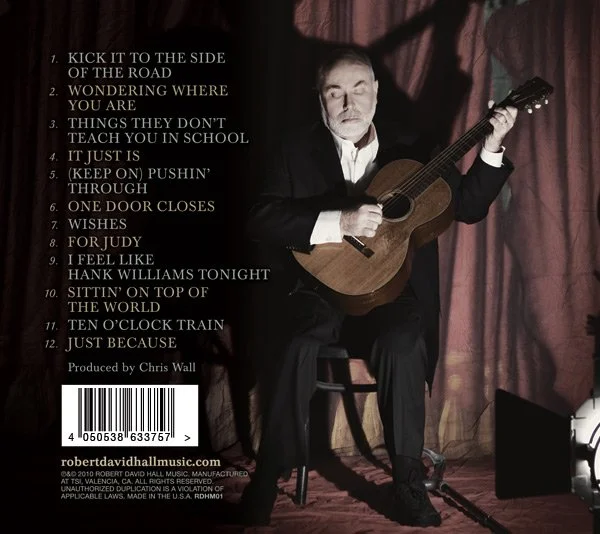 A man in a black suit sitting on a chair, playing an acoustic guitar in front of a red curtain background. Text on the left lists song titles and credits, with a barcode and website at the bottom.