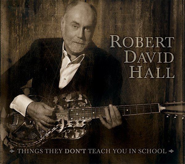 A man with a beard and mustache holding an acoustic guitar, dressed in a suit jacket and white shirt, sitting against a wooden wall. The text on the image reads 'Robert David Hall' and 'Things They Don't Teach You in School.'