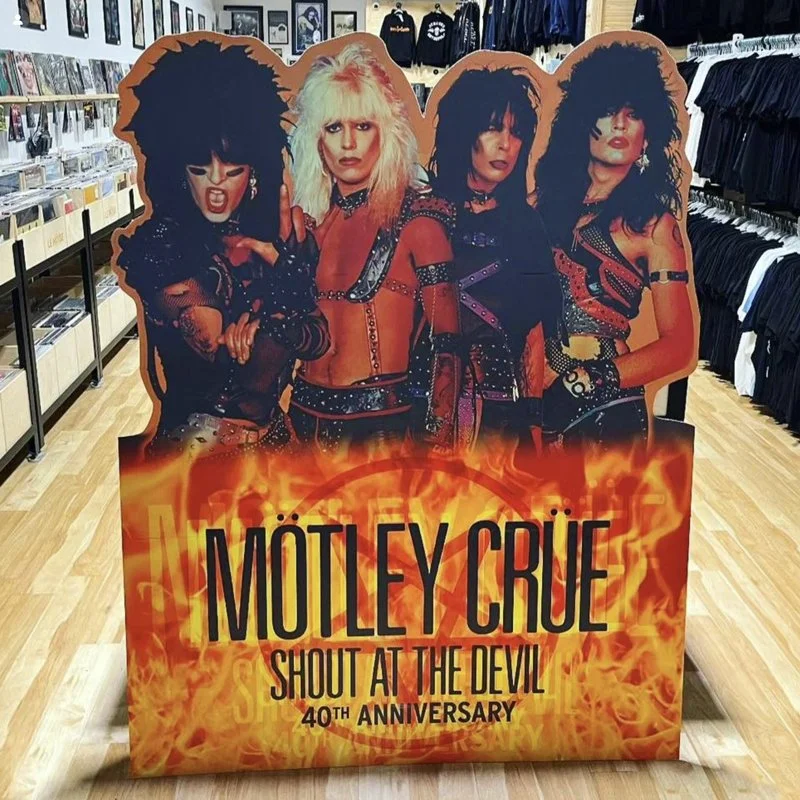 A cardboard cutout display of the band Mötley Crüe with four members dressed in 1980s glam metal style, surrounded by a fiery graphic, advertising their 40th anniversary of the album "Shout at the Devil" in a store aisle.