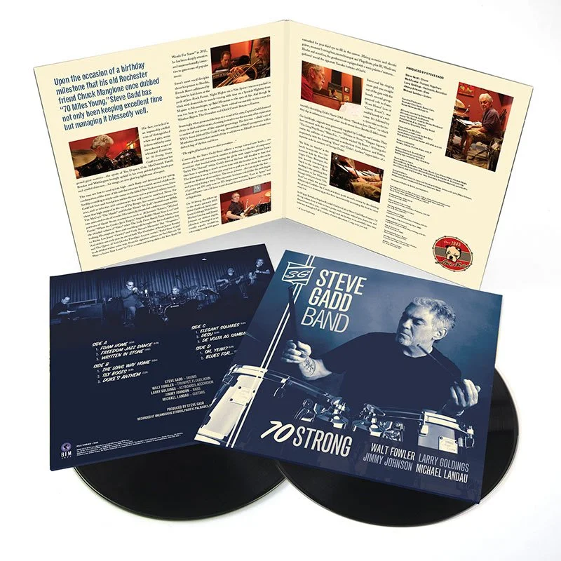 A vinyl record album titled "Steve Gadd Band - 70 Strong" by Steve Gadd Band with featured musicians Walt Fowler, Larry Goldings, Jimmy Johnson, and Michael Landau. The album cover shows Steve Gadd conducting with a baton, and the back cover contains the track list and band members. An open fold-out page features photographs and a written tribute to Steve Gadd.