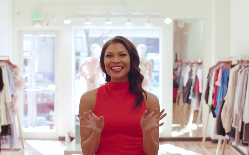 A woman in a red sleeveless dress inside a clothing store, smiling and gesturing with her hands.