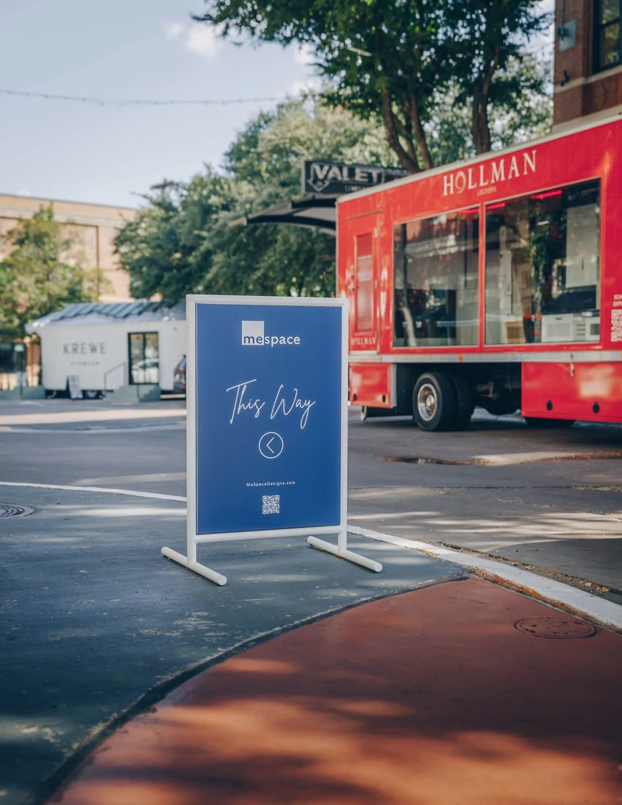 An outdoor sidewalk scene featuring a blue sign with white text that reads 'This Way' and an arrow pointing left, with a QR code below, next to a red food truck and a white kiosk in the background, under a partly cloudy sky with trees.