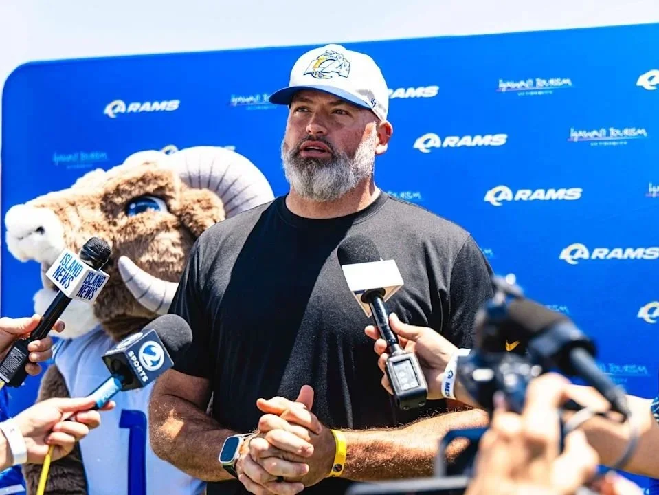 Man in a black T-shirt and white cap being interviewed by several reporters, with a Ram's mascot and blue branded backdrop in the background.